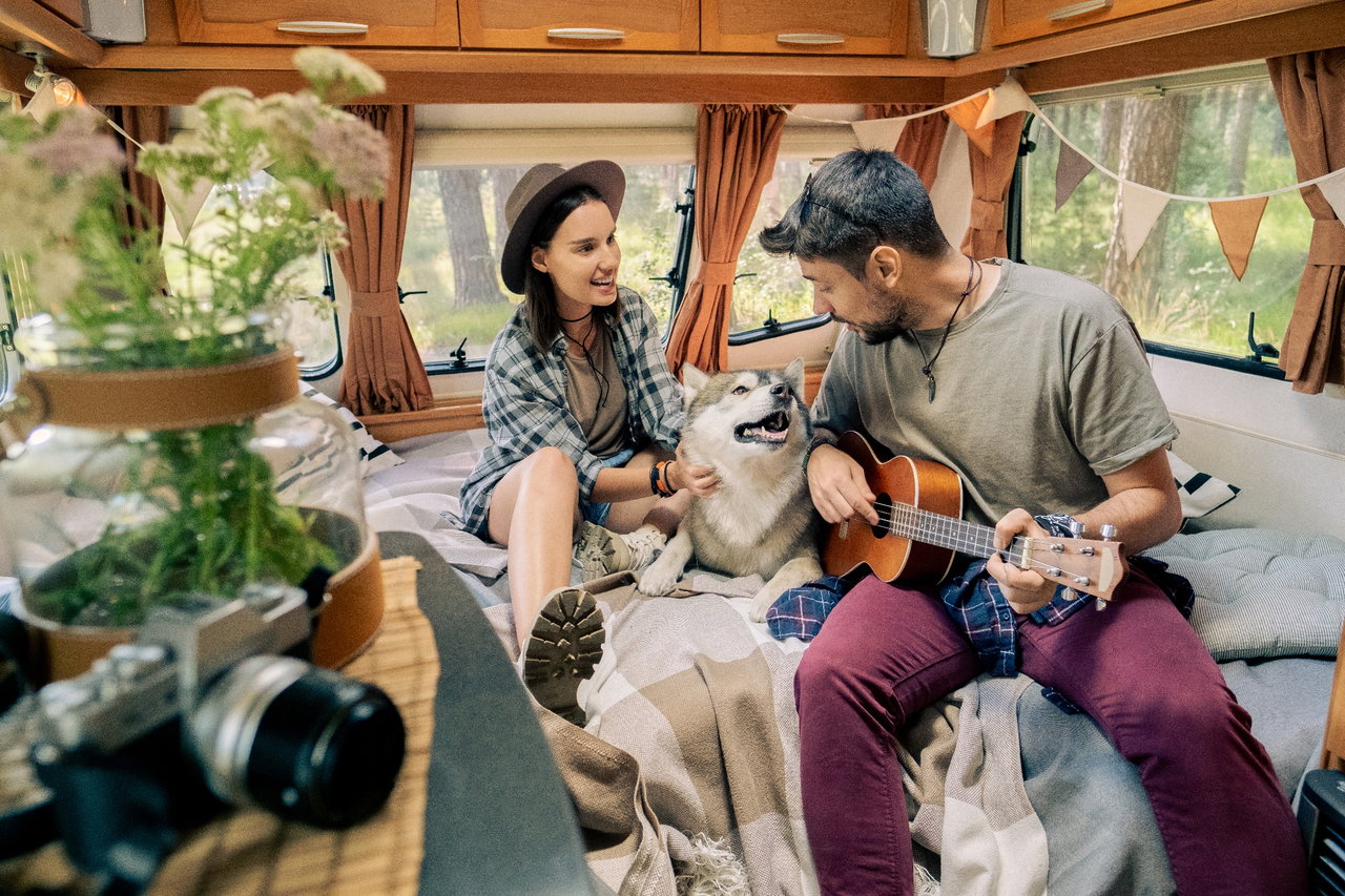 Paar sitzt in schön dekoriertem Campingfahrzeug mit ihrem Husky auf dem Bett, Mann spielt Gitarre.