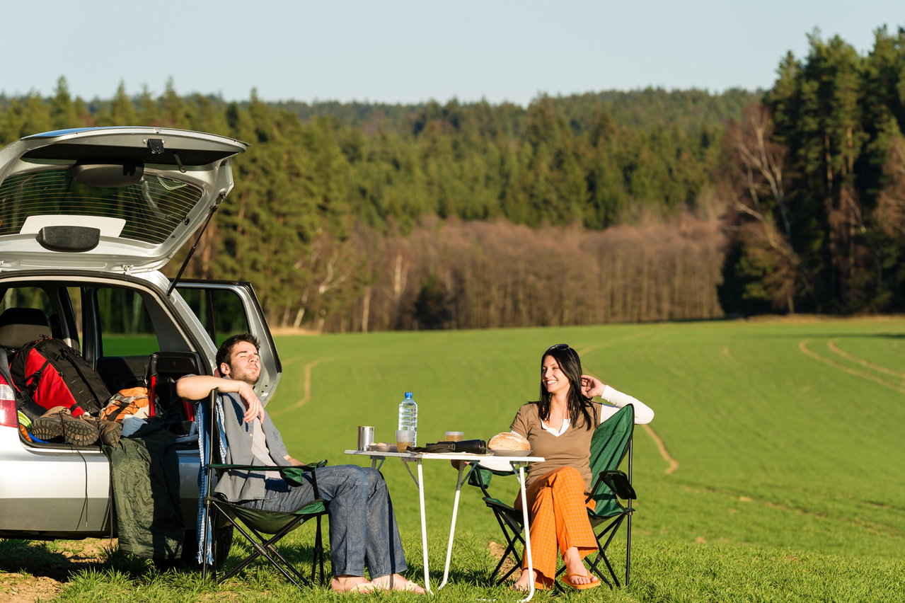 Ein Paar sitzt auf Campingstühlen in der Natur vor einem Auto mit geöffnetem Kofferraum.